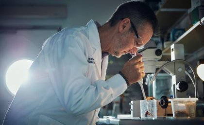 A male scientist in a lab coat and goggles holds a pipette above a large black spider in a container.